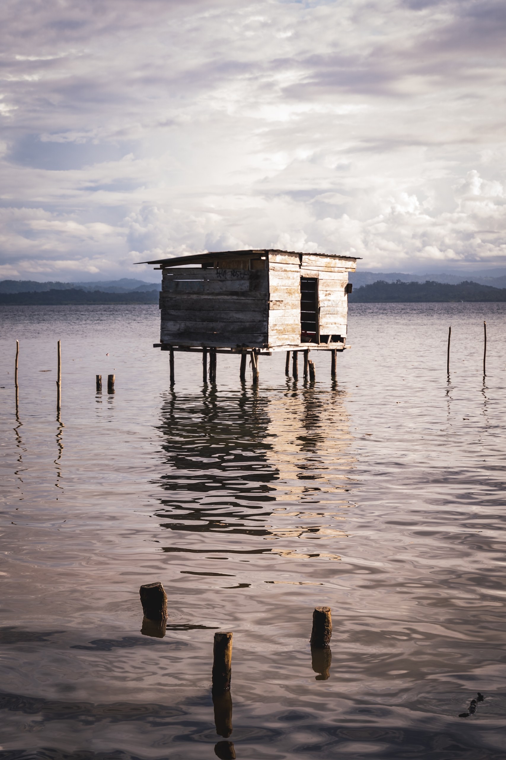 Weathered wooden hut on stilts standing in calm water, reflected on the surface, with old posts in the foreground and hills on the horizon, Santa Catalina, Panama