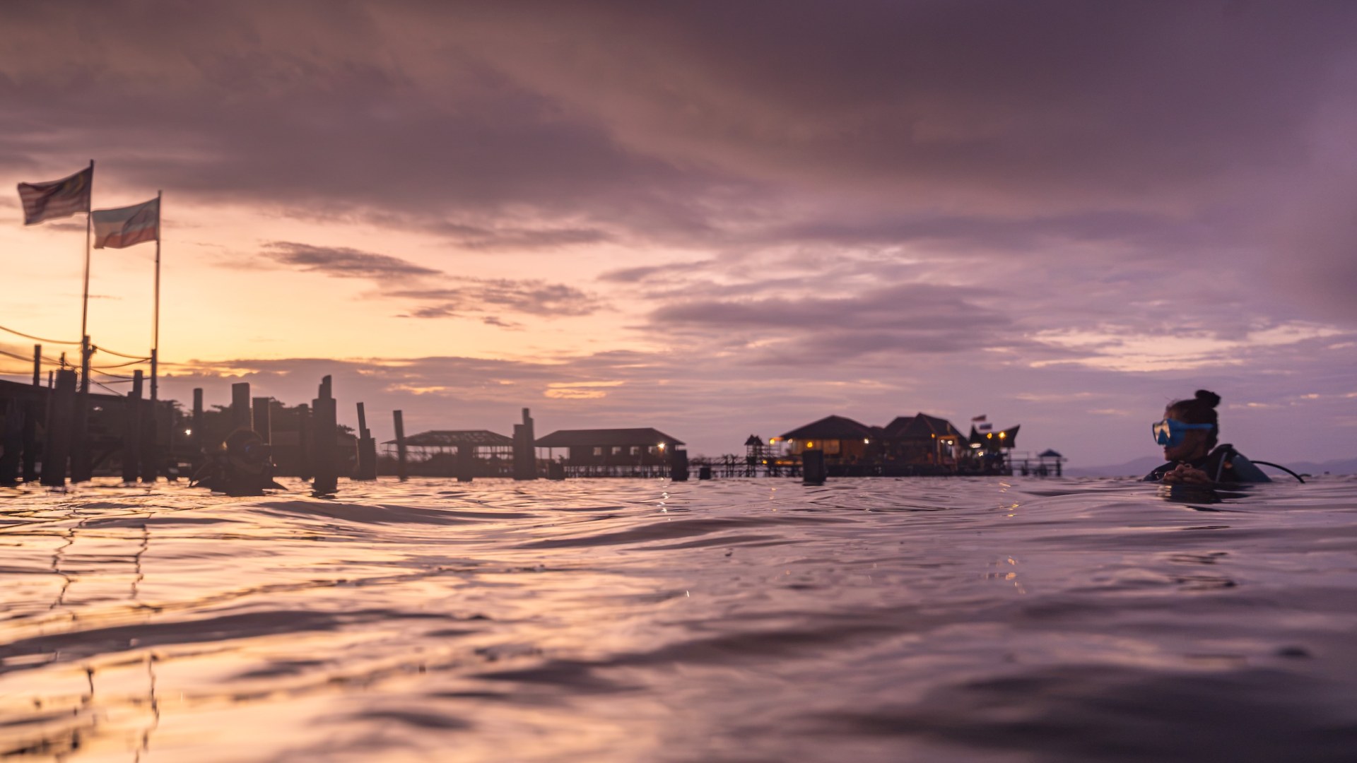 Scuba diver with a blue mask at the water's surface at dusk, stilt houses and flags in the background, Mabul, Malaysia