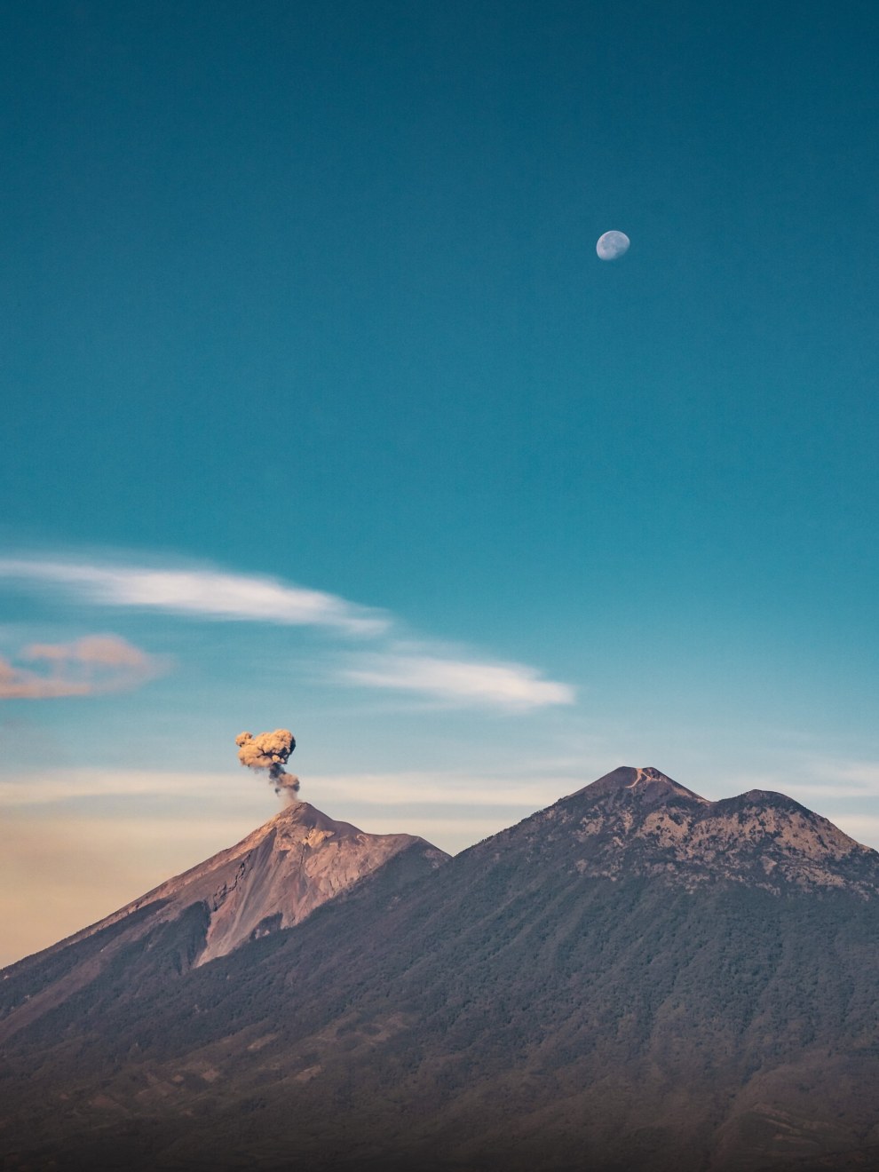 The erupting Volcán de Fuego emits a plume of ash, while the forested Volcán Acatenango rises to the right. The moon hangs in the blue evening sky above the volcanoes