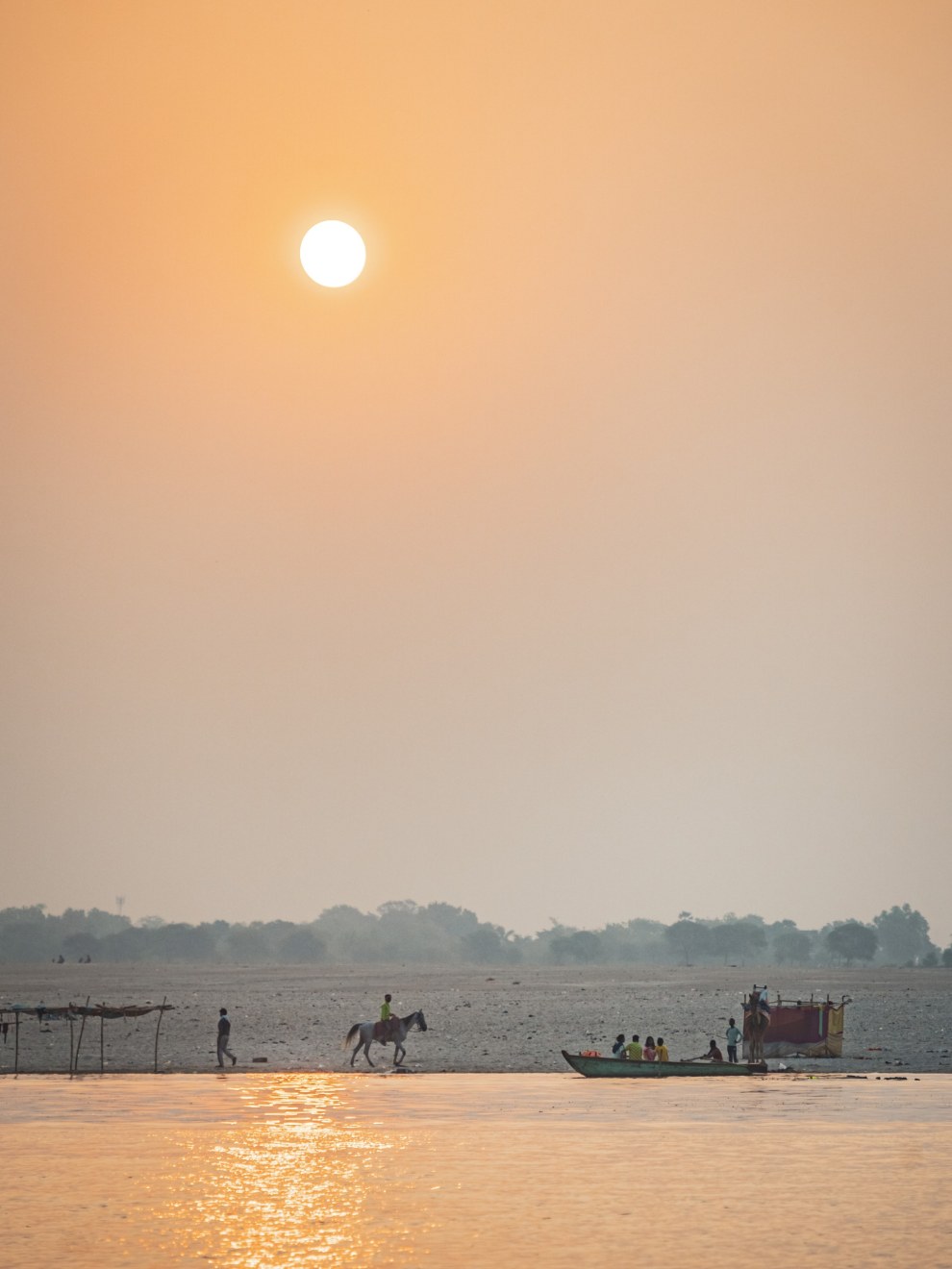 Hazy sunrise over the Ganges with a horse rider, people and a boat on the riverbank, Varanasi, India