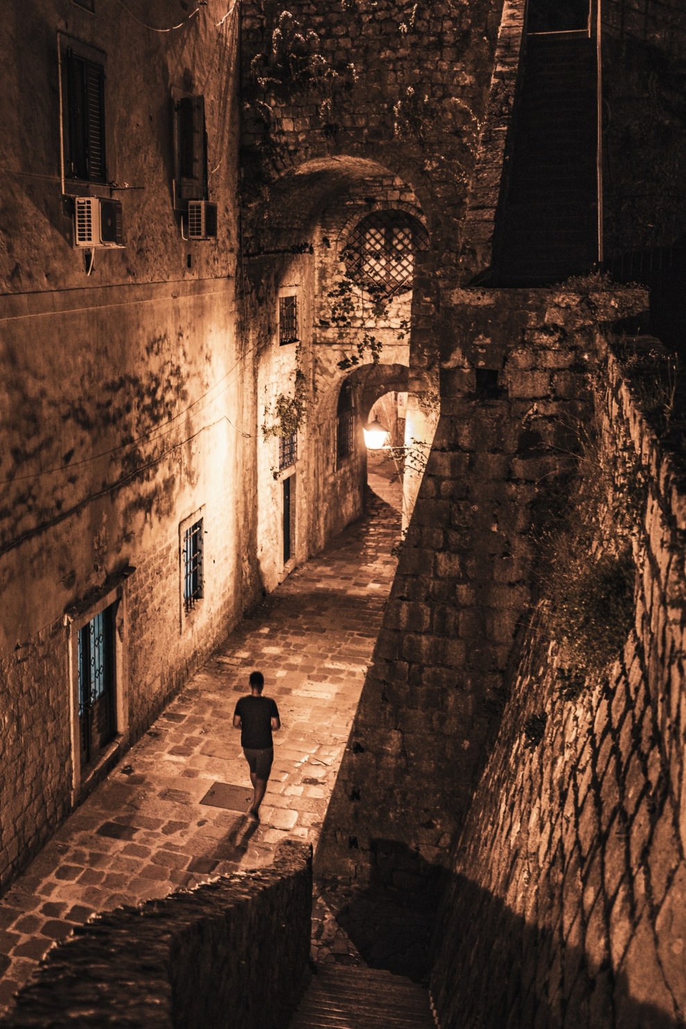 Person walking down a narrow stone alley lit by street lamps at night, Kotor, Montenegro