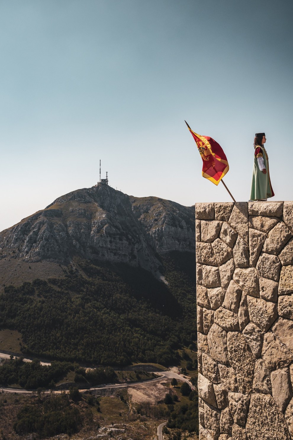 Person in historical dress beside a flag on a stone wall in front of a mountain at Lovćen, Montenegro