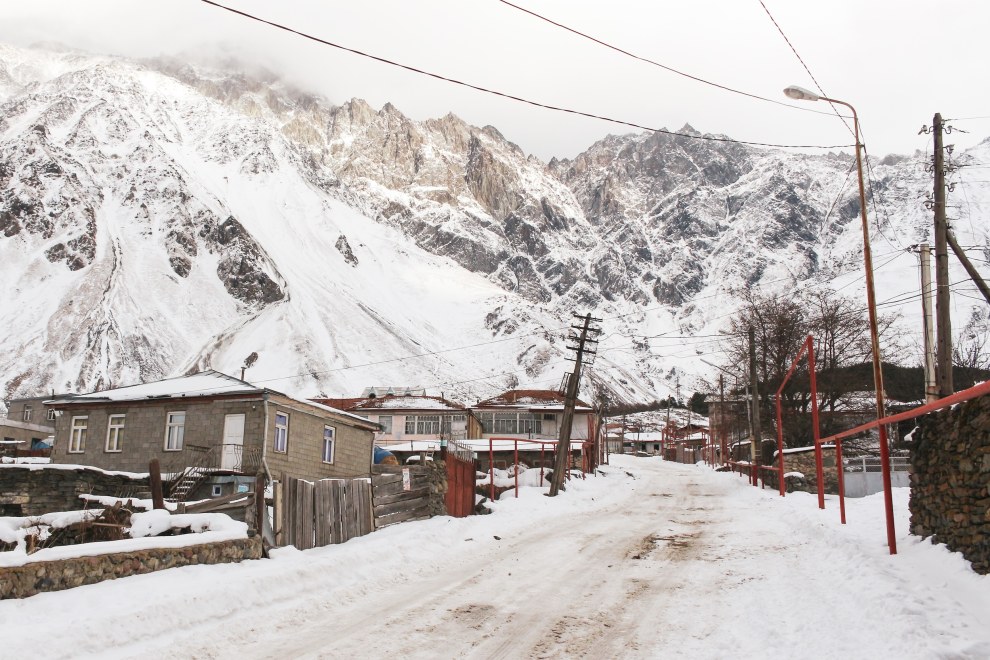 Snow-covered village street with houses, power lines and red fences below snowy mountains in Kazbegi, Georgia