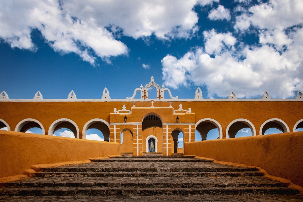 Yellow convent with arches and a stone staircase in Izamal, Mexico, under a blue sky with clouds
