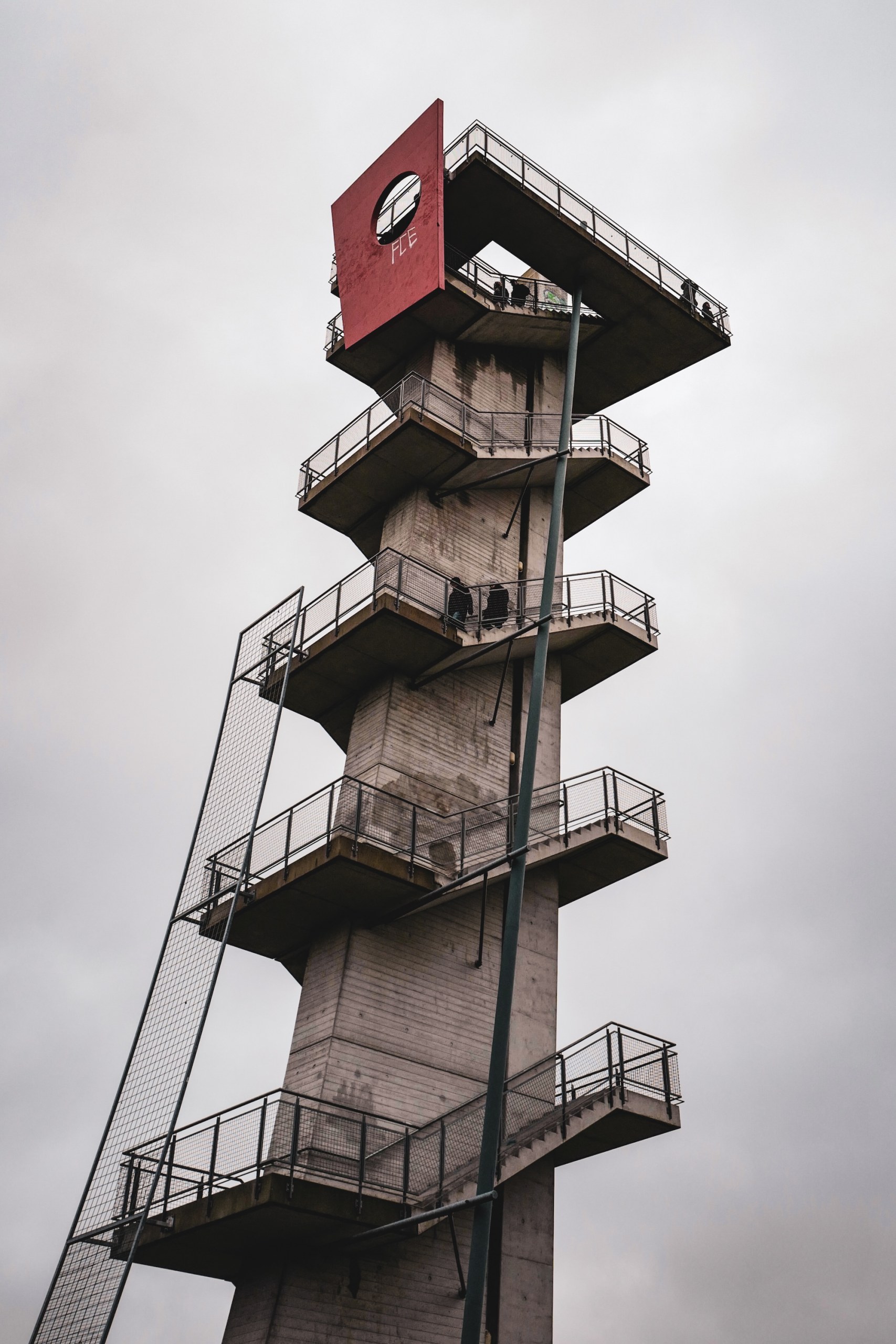 Merzdorf lookout tower near Cottbus: concrete tower with cantilevered stair platforms and a red top panel under a grey sky