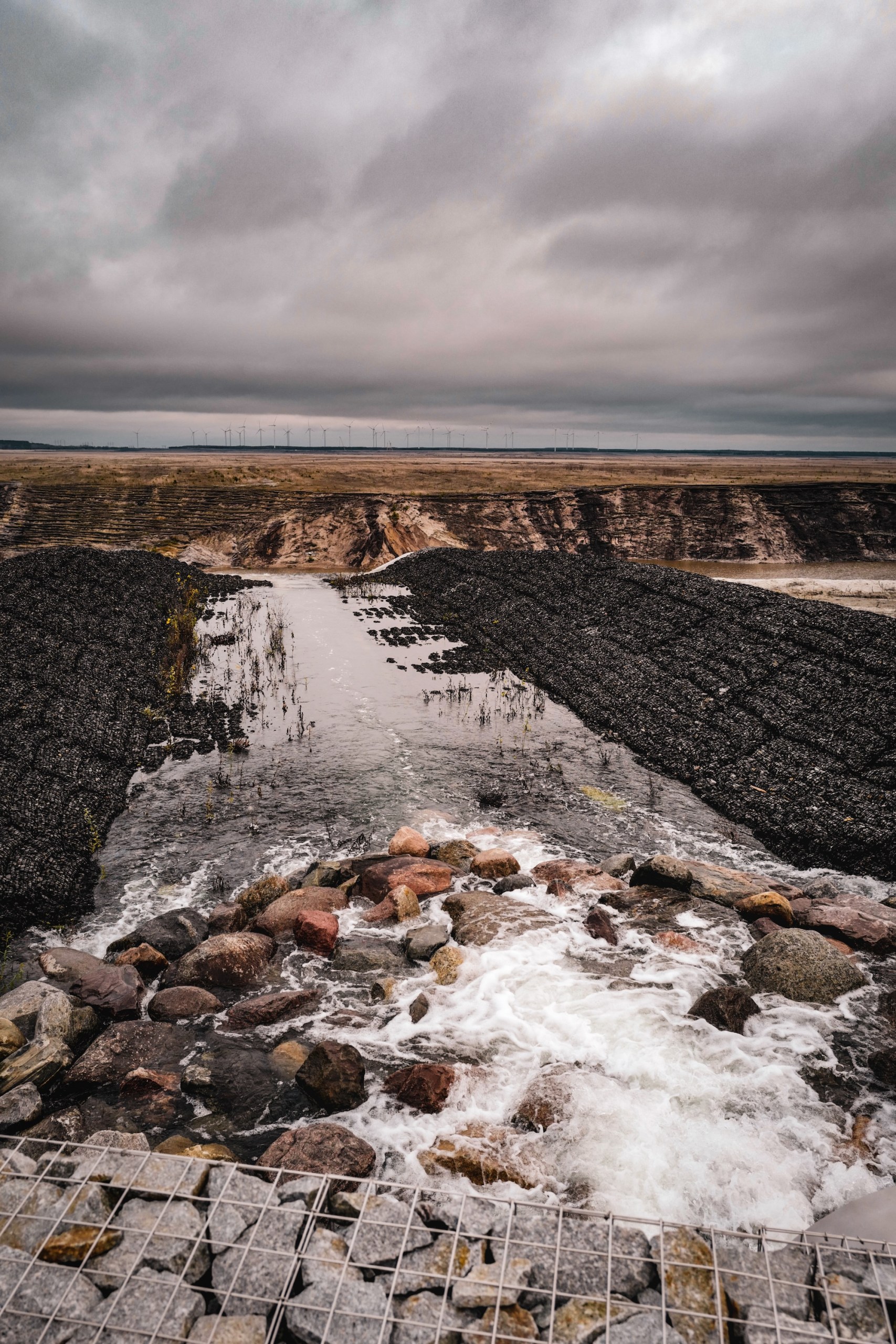 Water rushing over stones into the former open-cast mine that is being flooded to form the Cottbuser Ostsee, wind turbines on the horizon