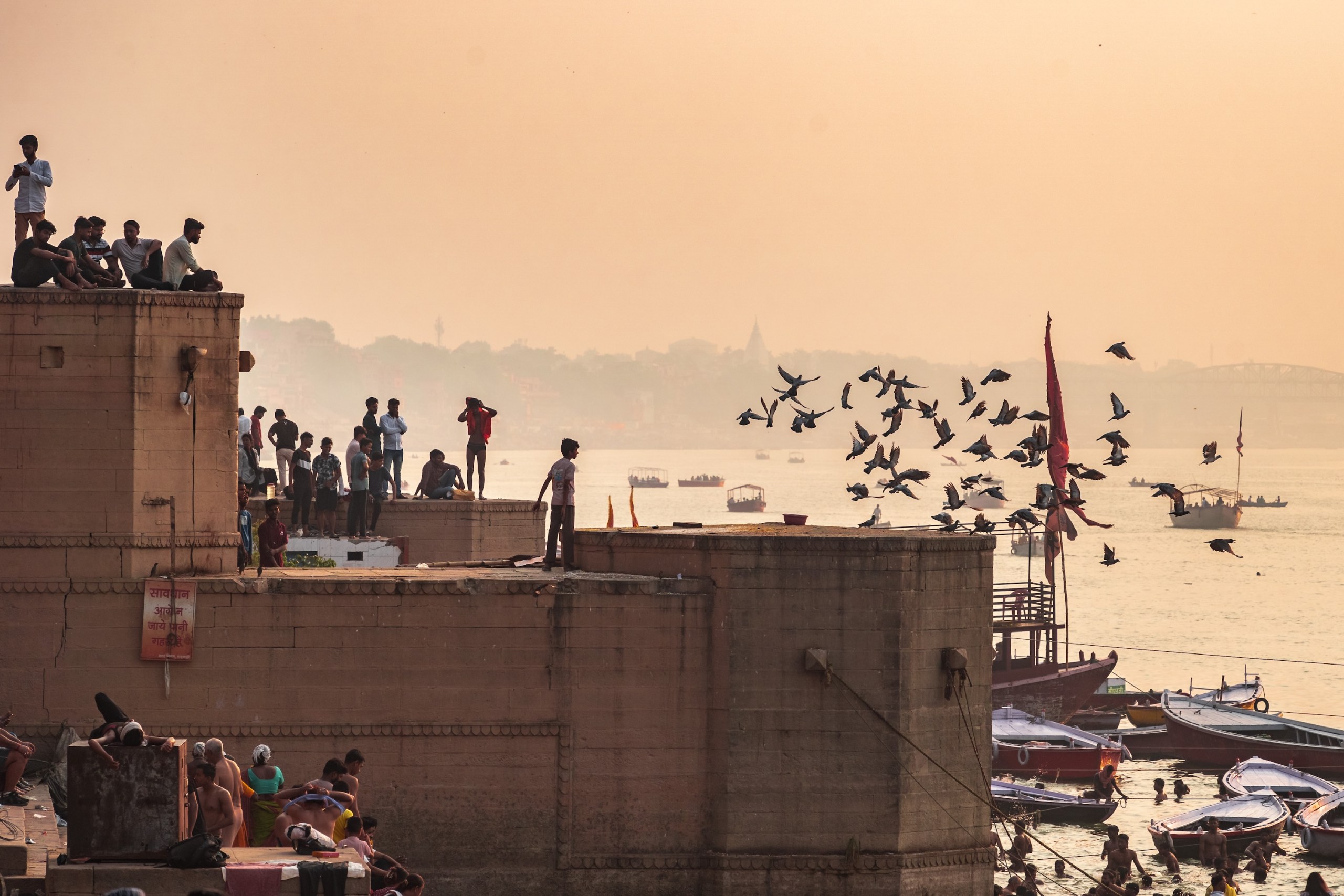 People on a ghat rooftop and a flock of birds above the Ganges in hazy golden light, Varanasi, India