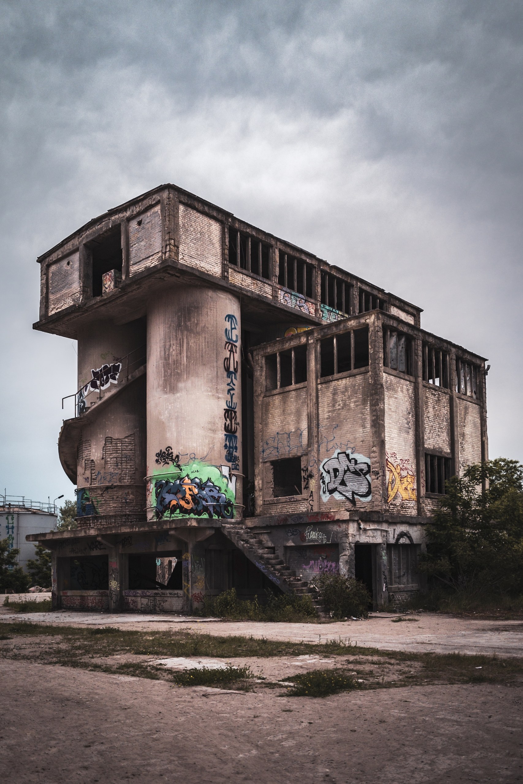 Derelict concrete industrial building with silo towers and graffiti under a cloudy sky