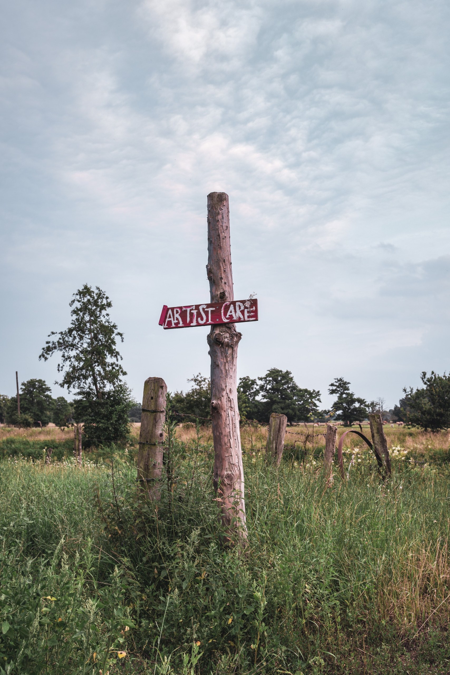 Wooden post with a hand-painted "Artist Care" sign at the edge of a meadow on the deserted MOYN festival site, near Oyten, Germany, summer 2020