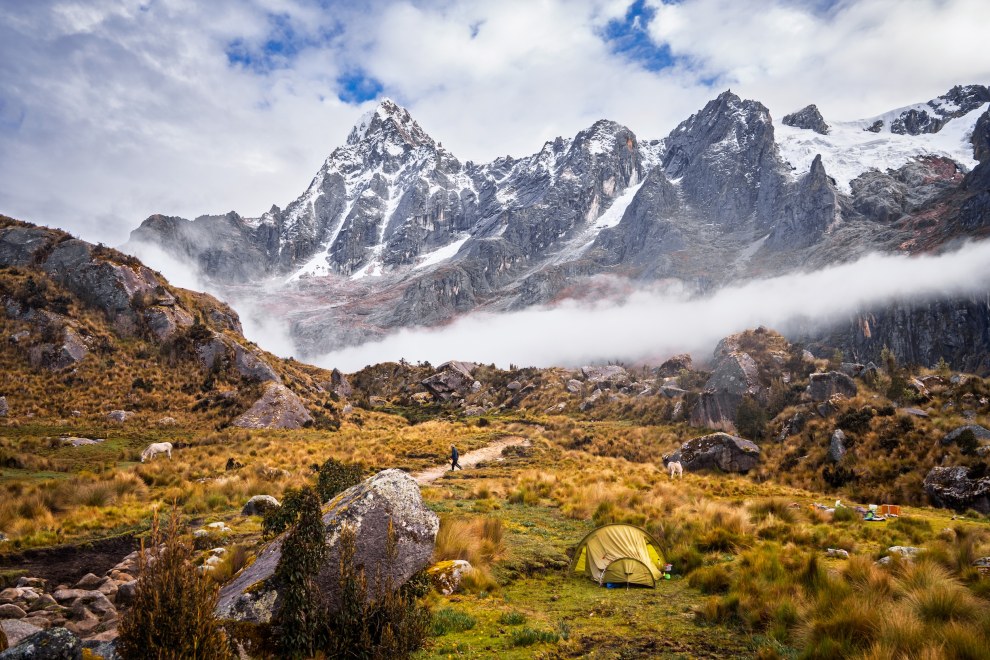 Small tent in a high mountain meadow beneath jagged snow-covered peaks and a band of clouds, Cordillera Blanca, Peru