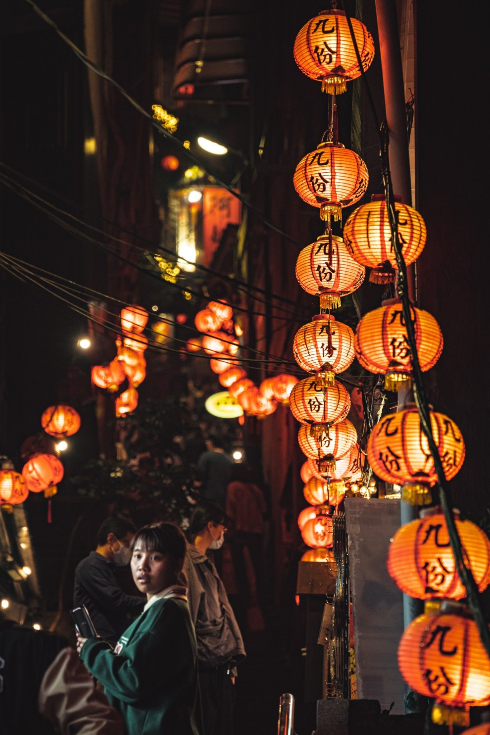 Red paper lanterns glowing above a narrow alley at night with a young woman looking back, Jiufen, Taiwan