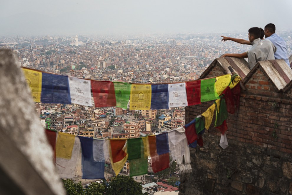 Prayer flags in front of a view over the densely built Kathmandu valley as two men point into the distance, Nepal