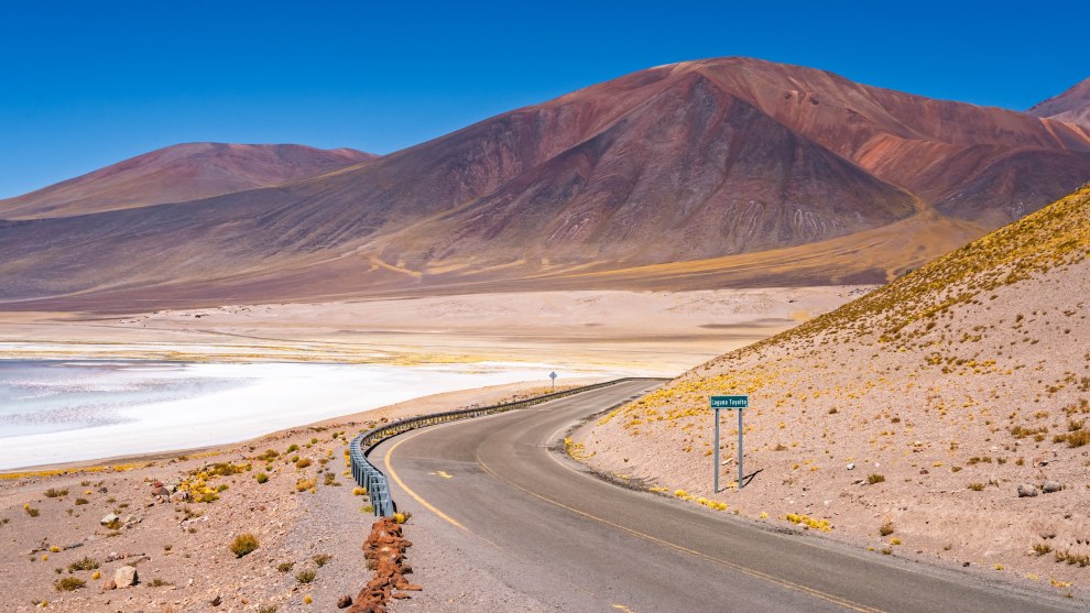 Road curving past the salt lake Laguna Tuyajto beneath reddish volcanic mountains in the Atacama, Chile