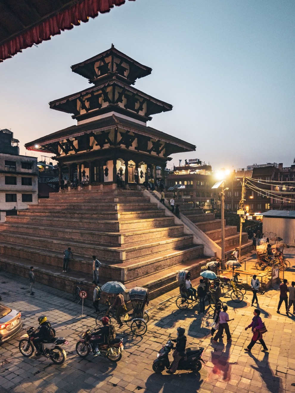 Tiered Maju Deval temple on its brick plinth at dusk with rickshaws and motorbikes casting long shadows, Kathmandu, Nepal