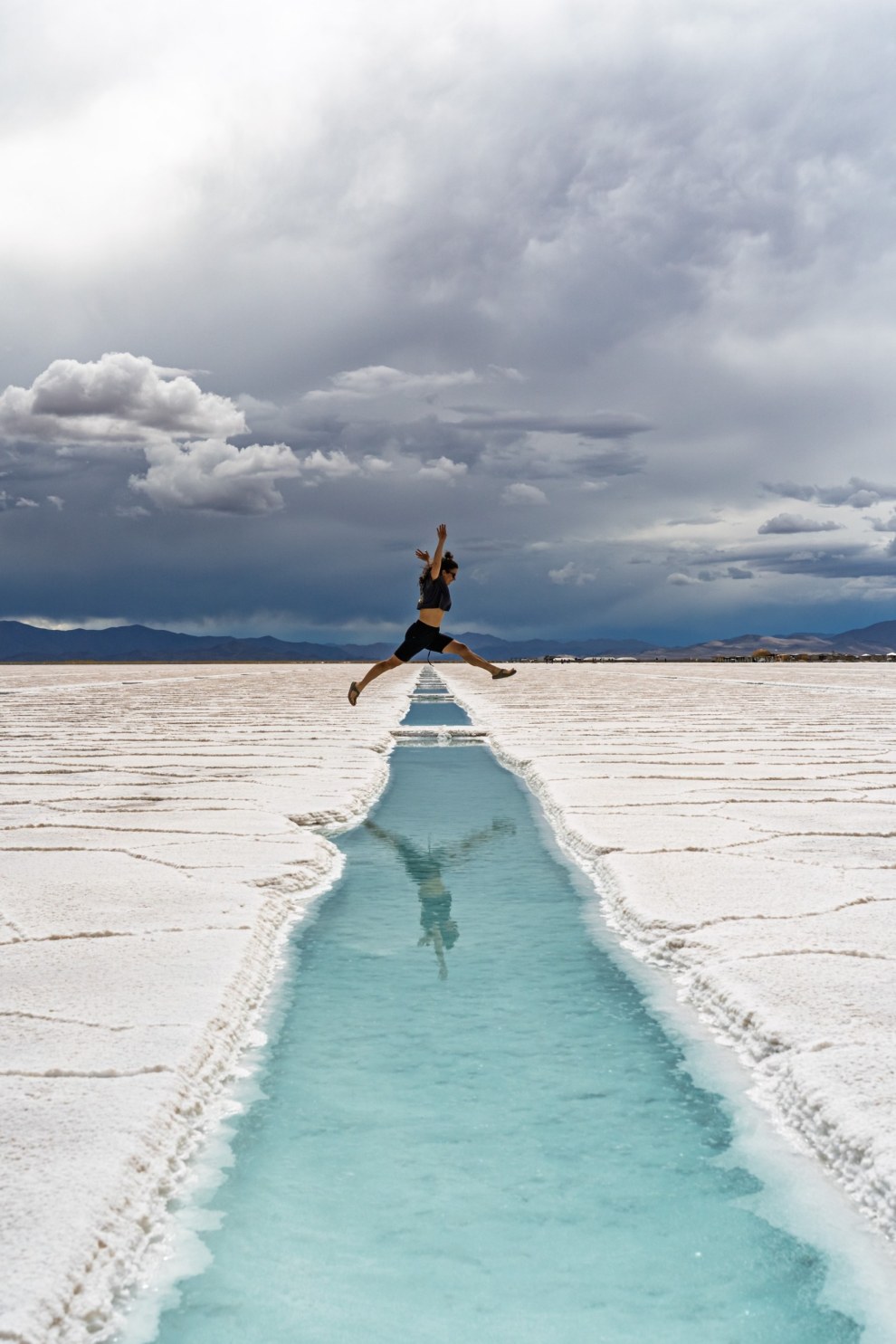 Person leaping across a turquoise water channel cut into the white salt flats of Salinas Grandes, Argentina