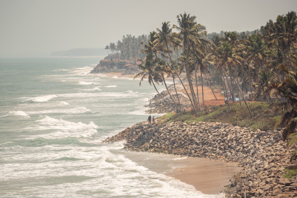 Waves breaking on a rocky coastline lined with palm trees in the haze, Varkala, India