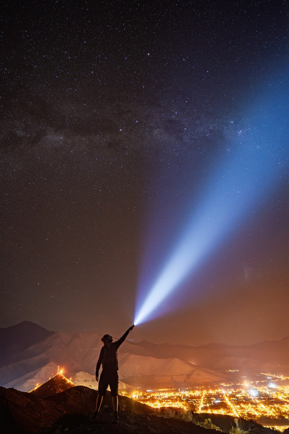 Silhouette of a man pointing a flashlight beam into the Milky Way above the lights of Vicuña, Chile