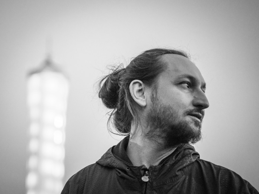 Black-and-white portrait of photographer Danilo Rößger in profile, with a blurred tower in the background, Erlebnispark Teichland, Cottbus, Germany