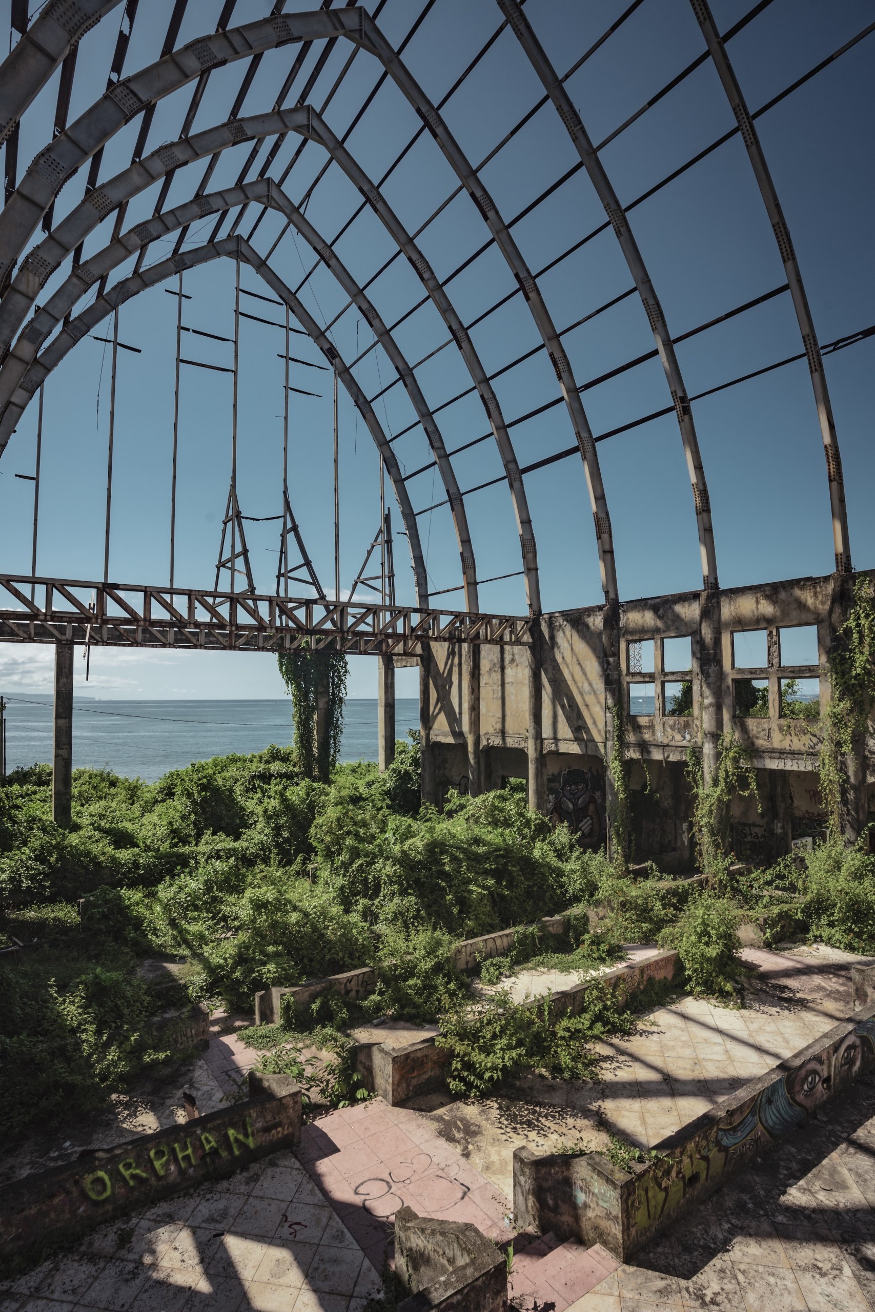 Rusted arched skeleton of a greenhouse above overgrown ruins with the sea behind, Taman Festival, Bali, Indonesia