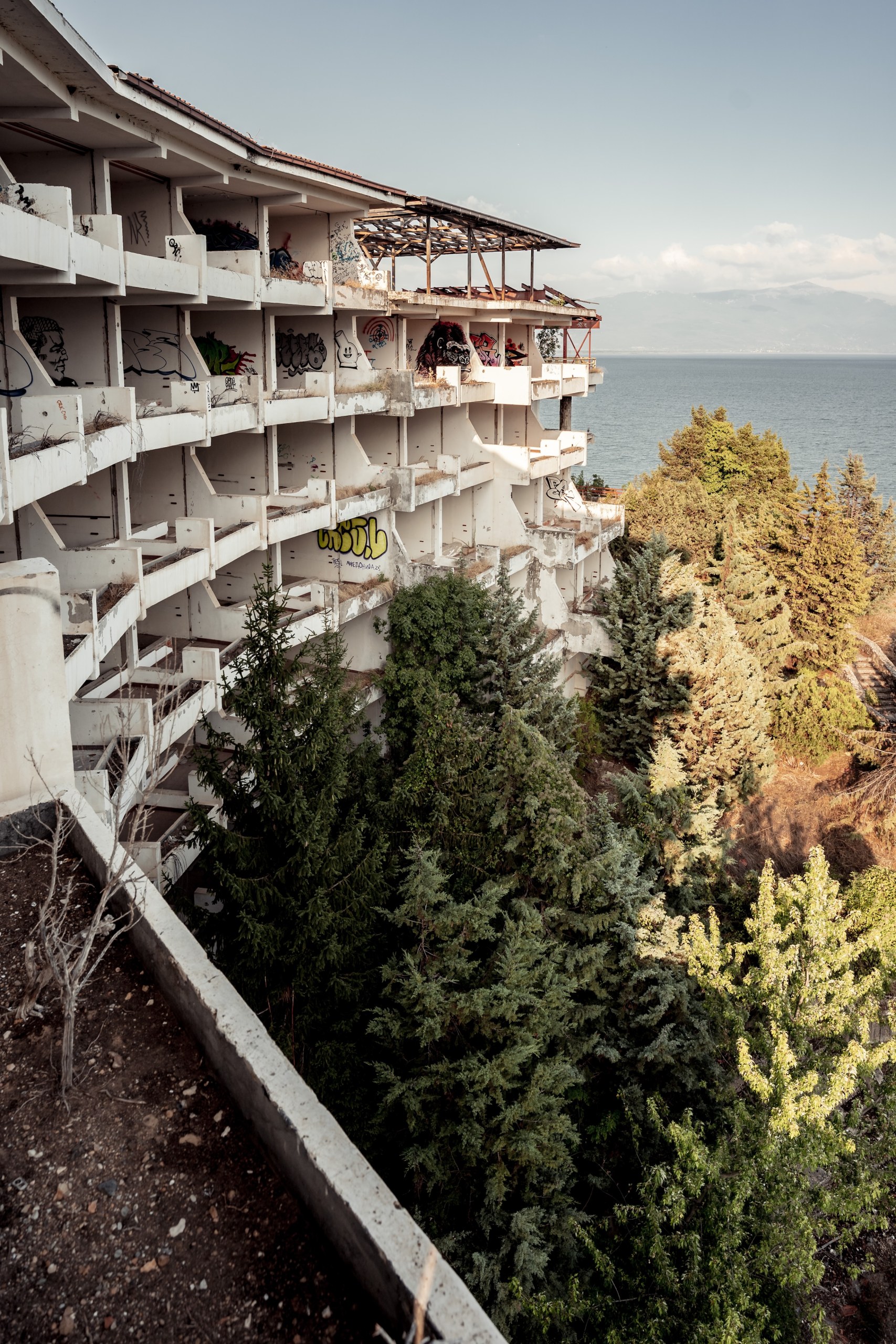 Abandoned hotel with graffiti-covered balconies overgrown by trees above Lake Prespa, North Macedonia