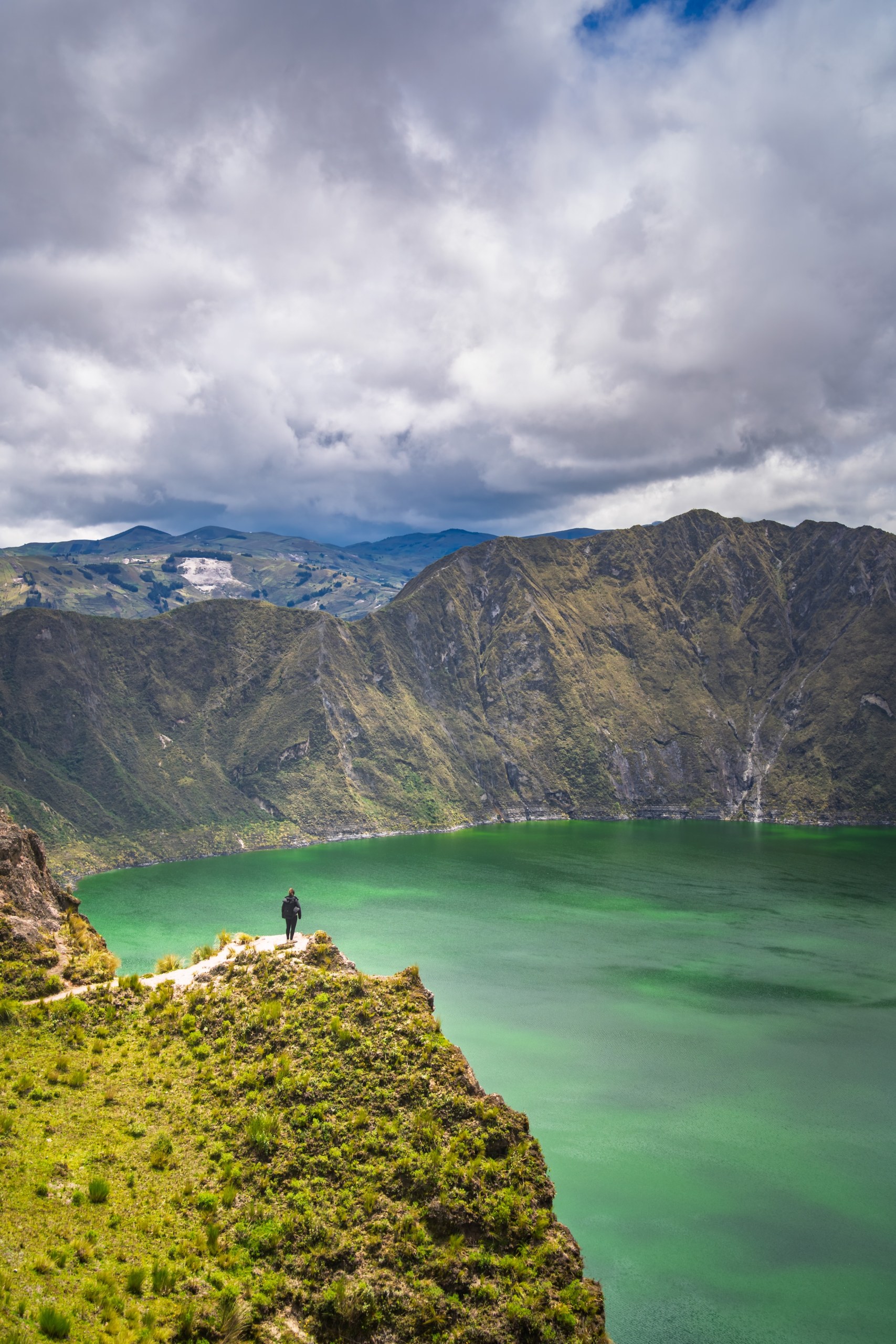 Person standing on a grassy ledge above the green crater lake of Quilotoa under heavy clouds, Ecuador