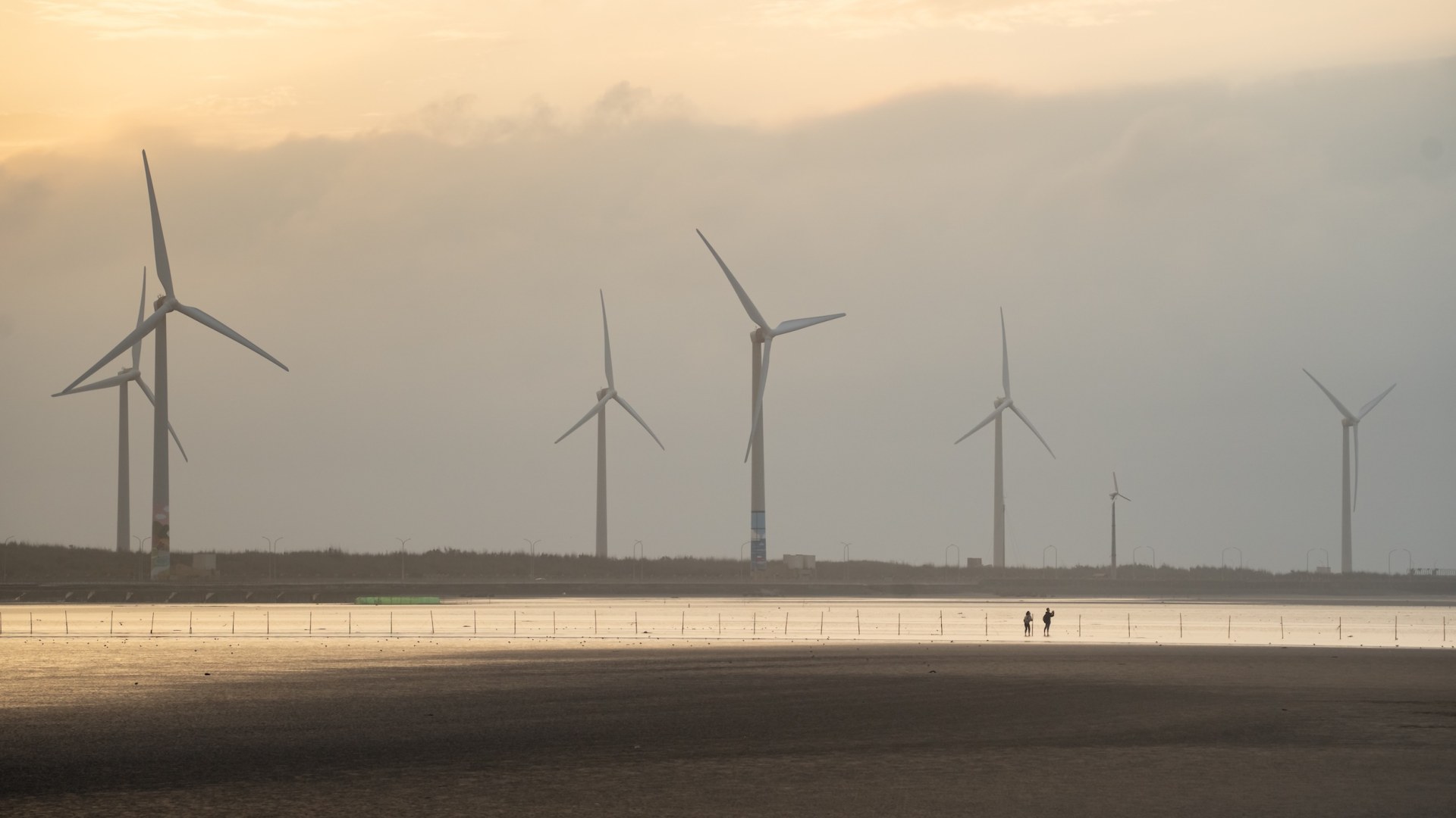 Two people on the tidal flats beneath a row of wind turbines in hazy evening light, Gaomei, Taiwan