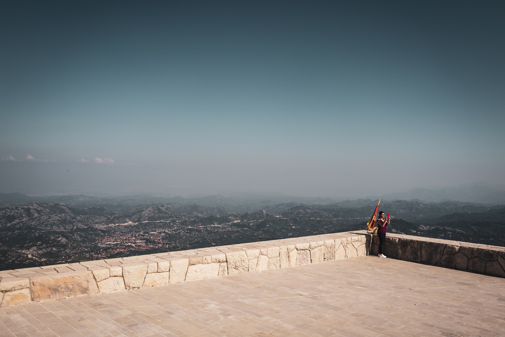 Woman photographing the view beside a flag on a stone viewing terrace high above a hazy landscape, Lovćen, Montenegro