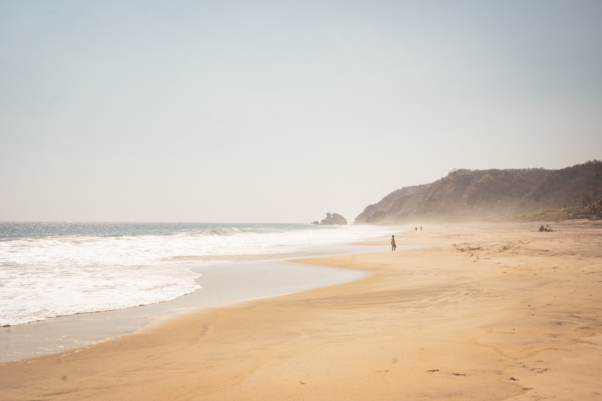 Lone figure walking along a wide, hazy beach with surf and a cliff in the distance, Varkala, India