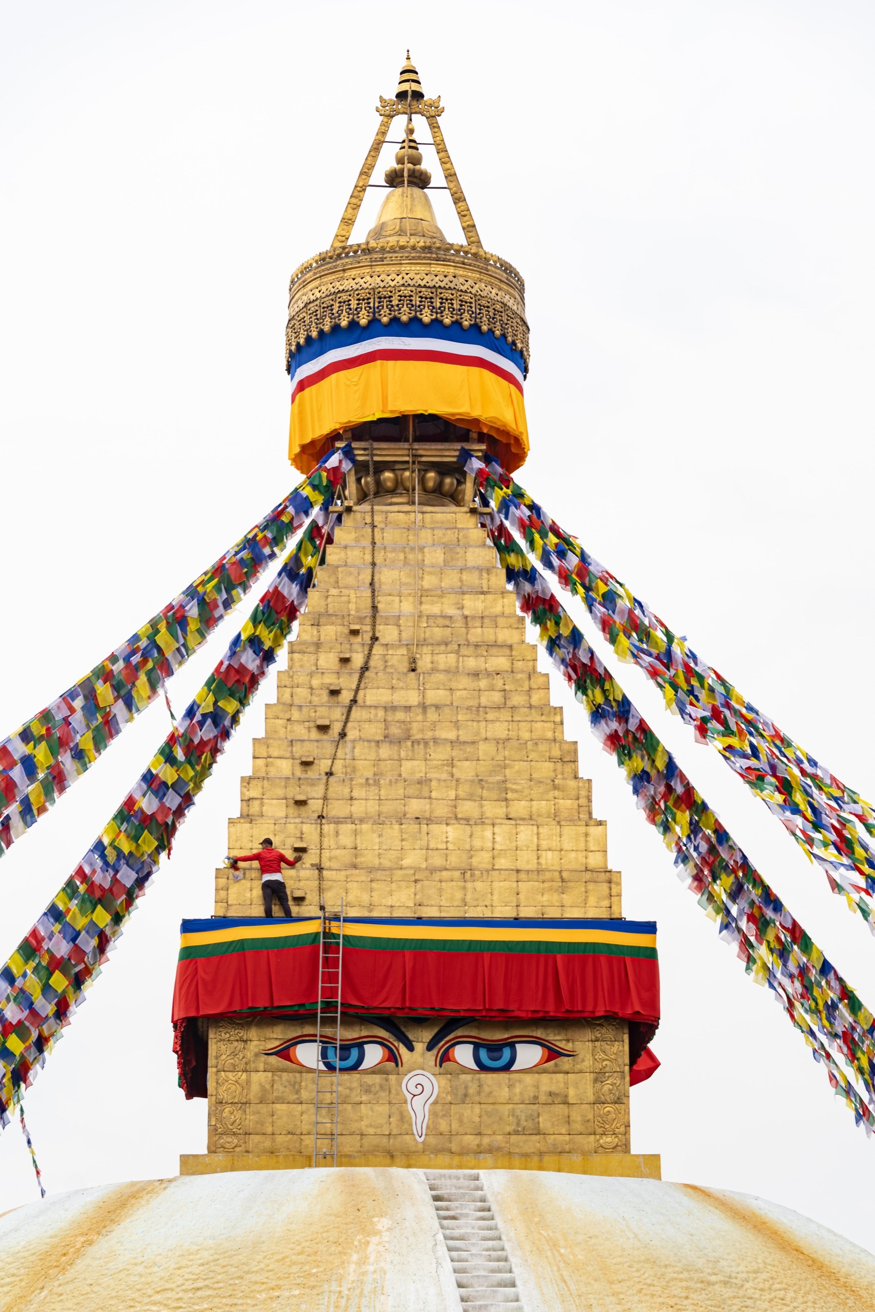 Worker on the golden spire of Boudhanath Stupa beneath painted eyes and strings of prayer flags, Kathmandu, Nepal