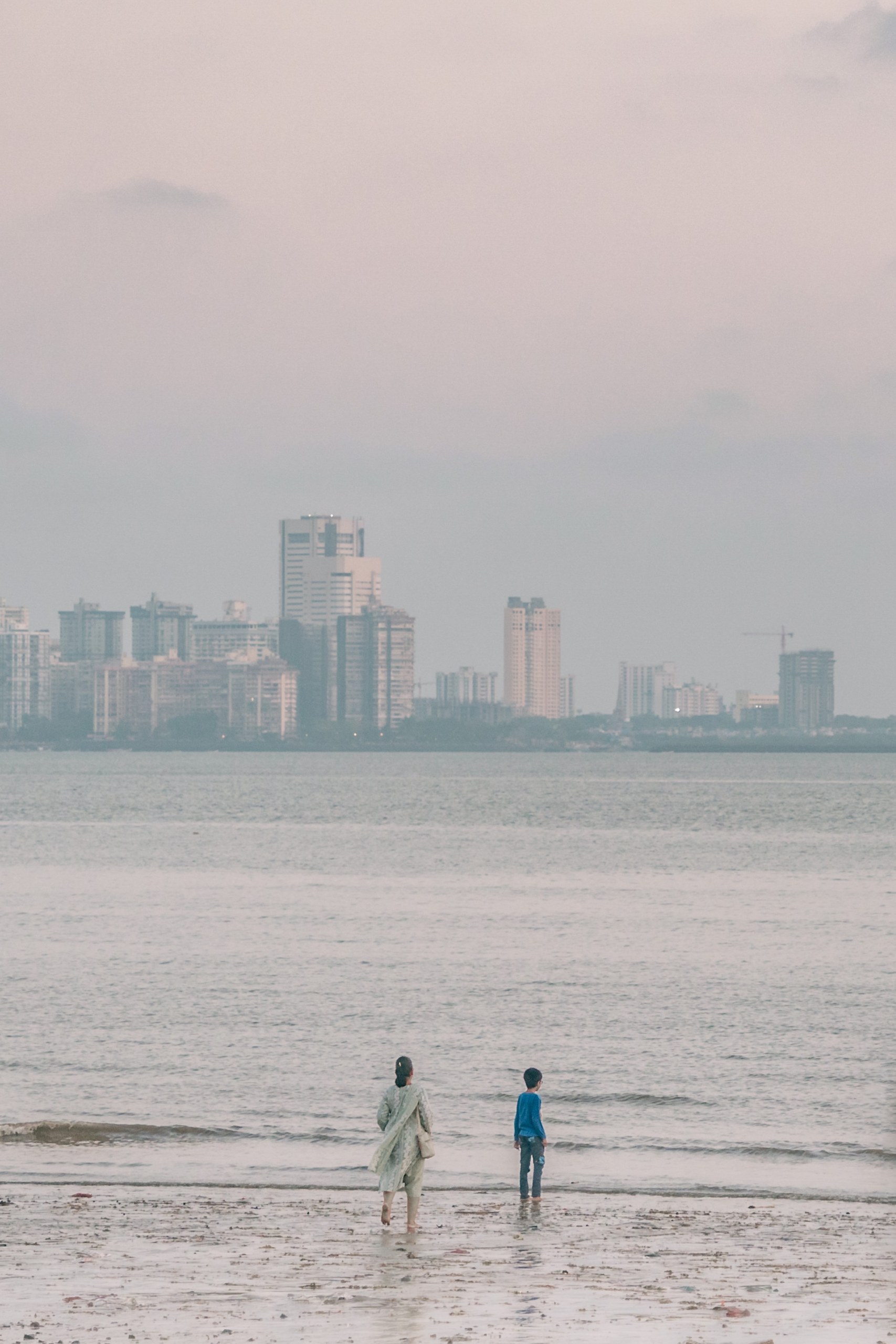 Woman and child at the water's edge in front of the hazy skyline of Mumbai, India