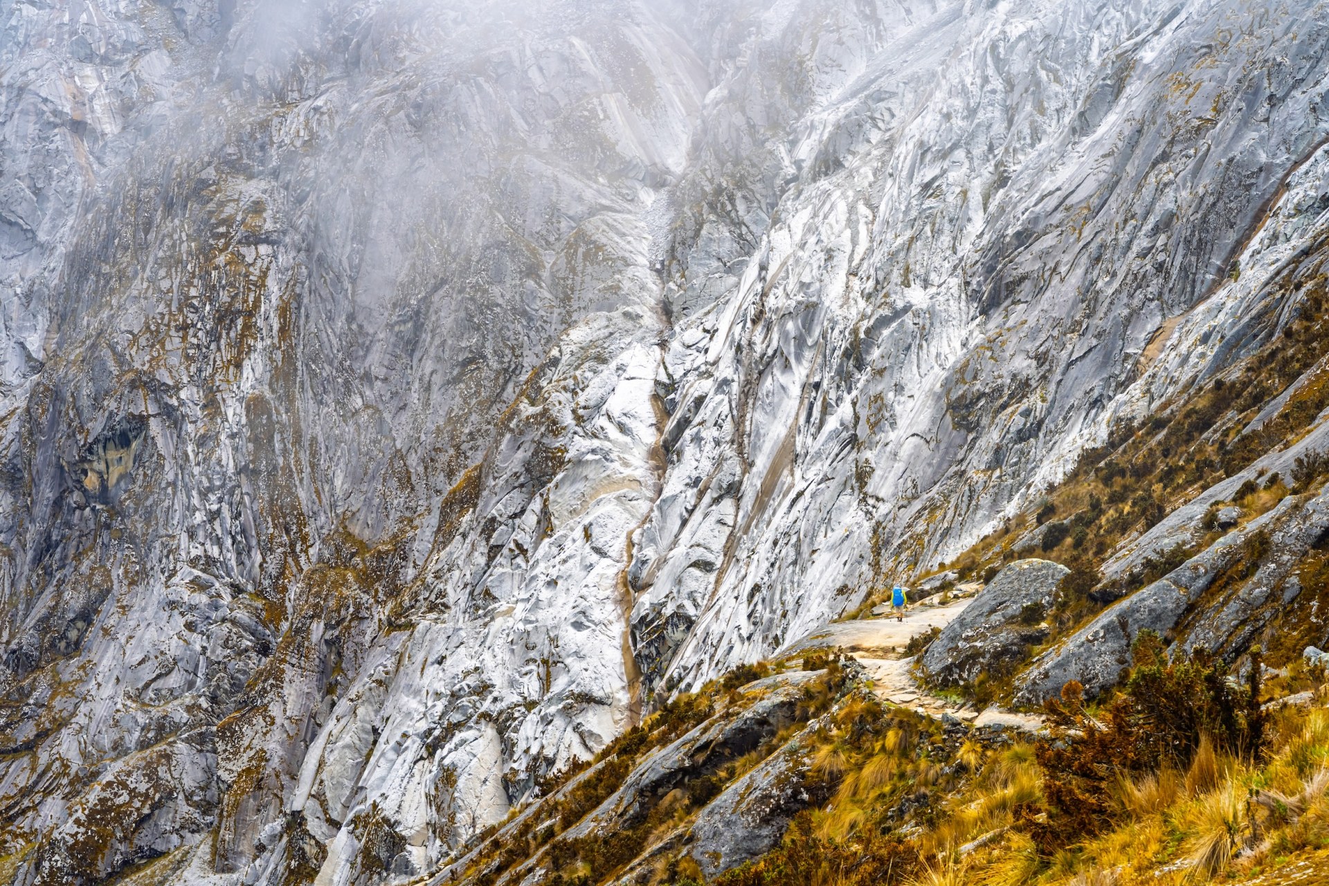 Hiker with a blue backpack on a trail beneath a vast, mist-covered granite wall in the Cordillera Blanca, Peru