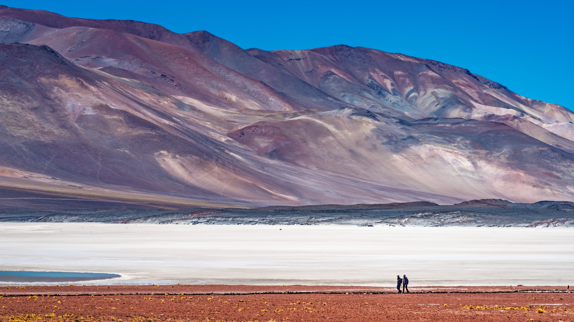 Two hikers walking past a white salt flat beneath colourful volcanic slopes at Piedras Rojas, Chile