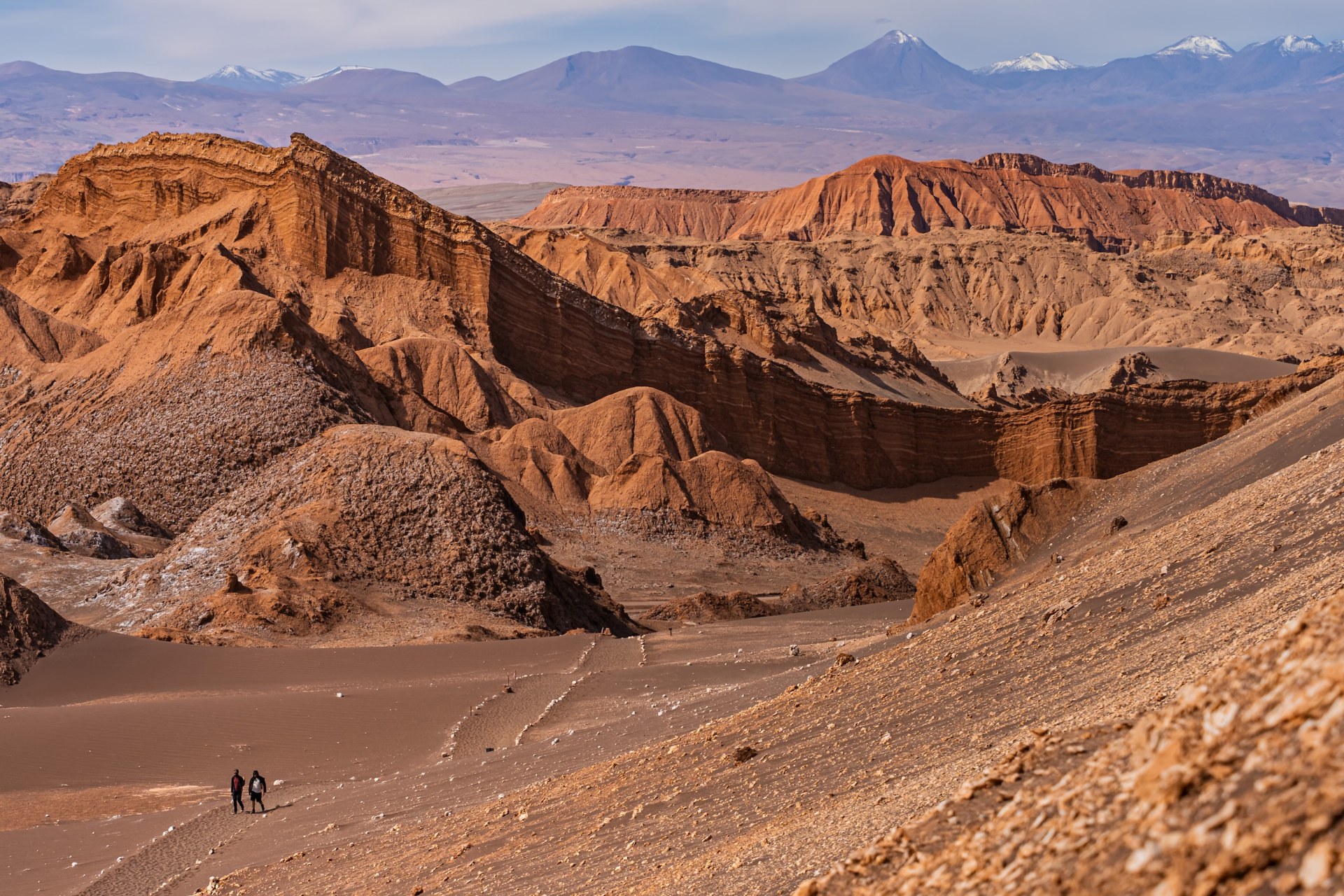 Two hikers on a sandy trail among towering eroded rock formations in the Valle de la Luna, Chile