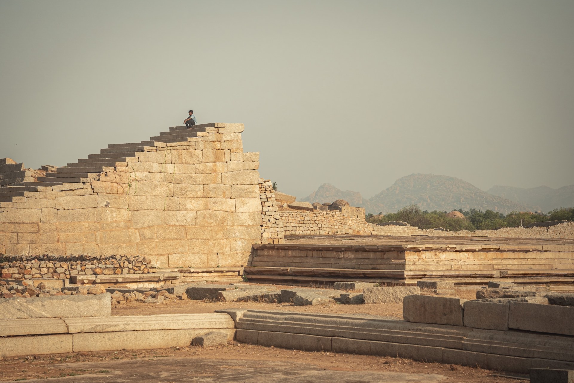 Boy sitting on top of a stepped stone platform among ancient ruins, Hampi, India