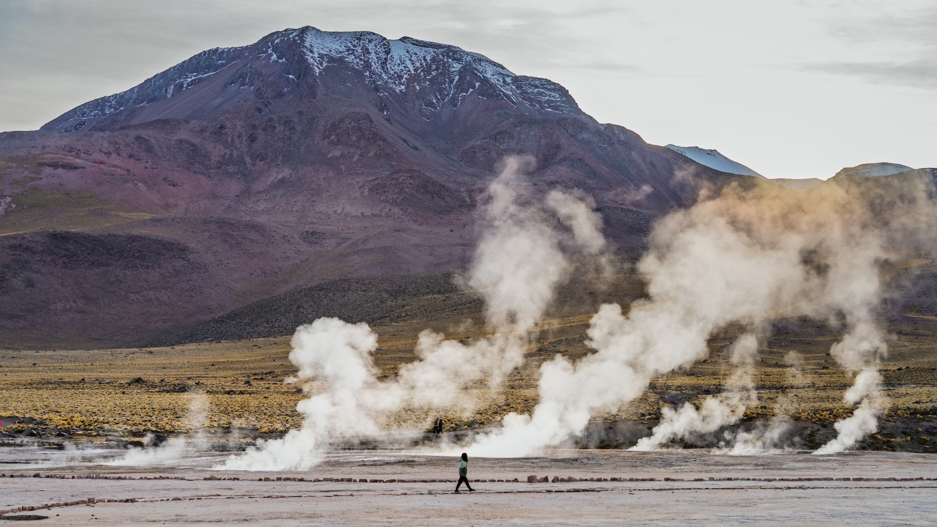 Person walking past steaming geysers beneath a snow-dusted mountain at El Tatio, Chile
