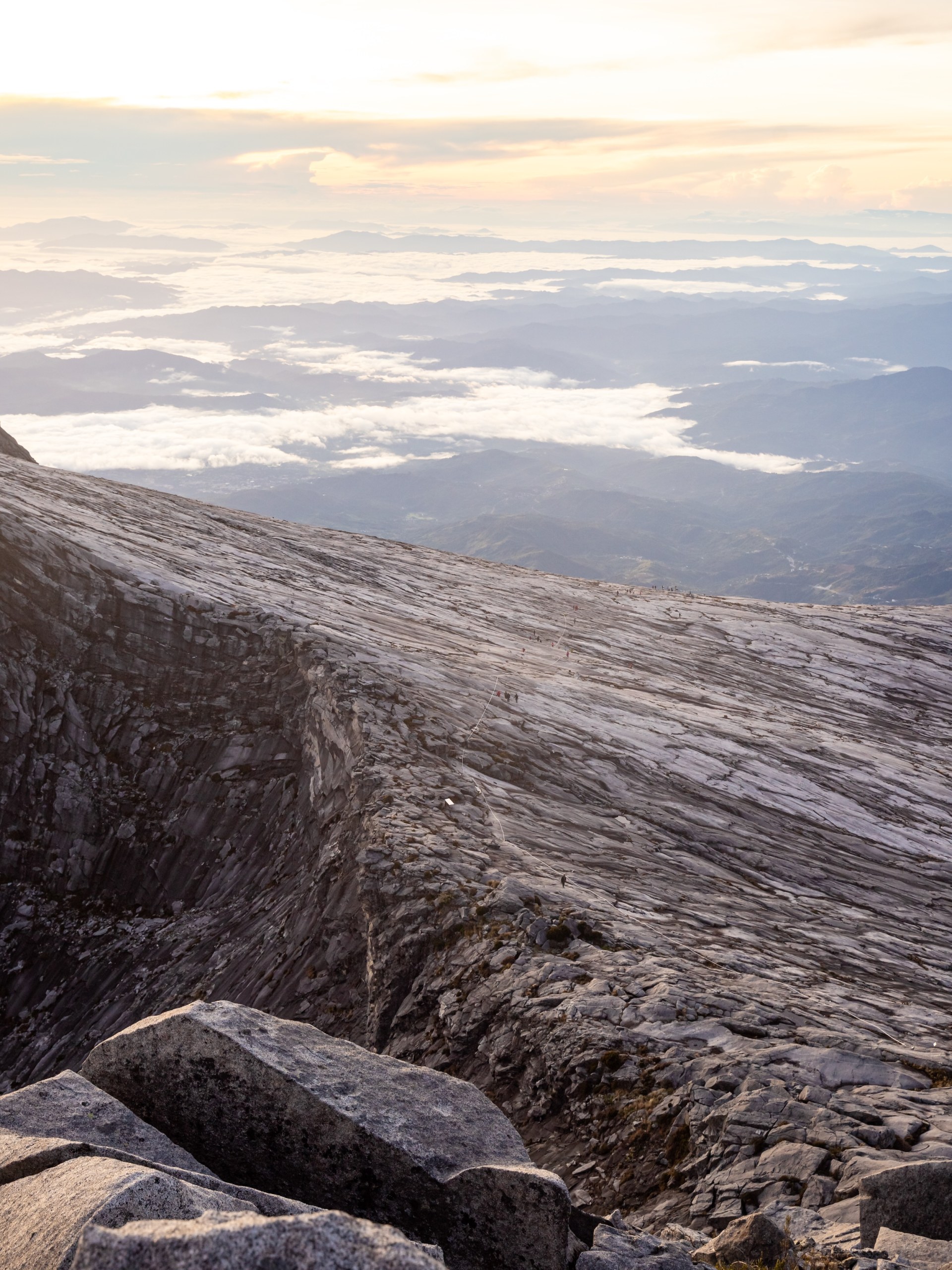Tiny climbers following a rope across the vast granite summit plateau of Mount Kinabalu above a sea of clouds at sunrise, Malaysia