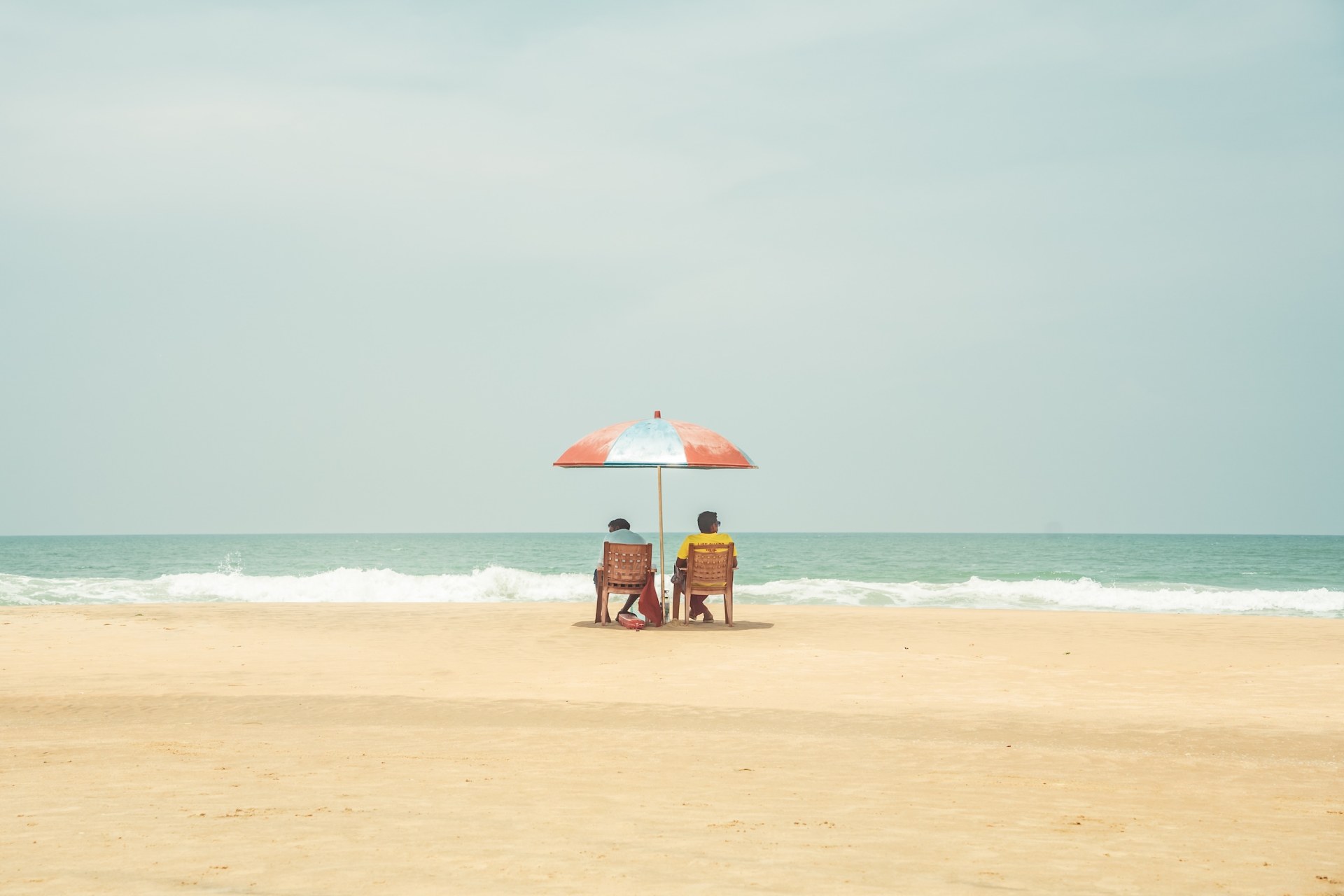 Two people sitting under a beach umbrella on an empty sandy beach in Varkala, India