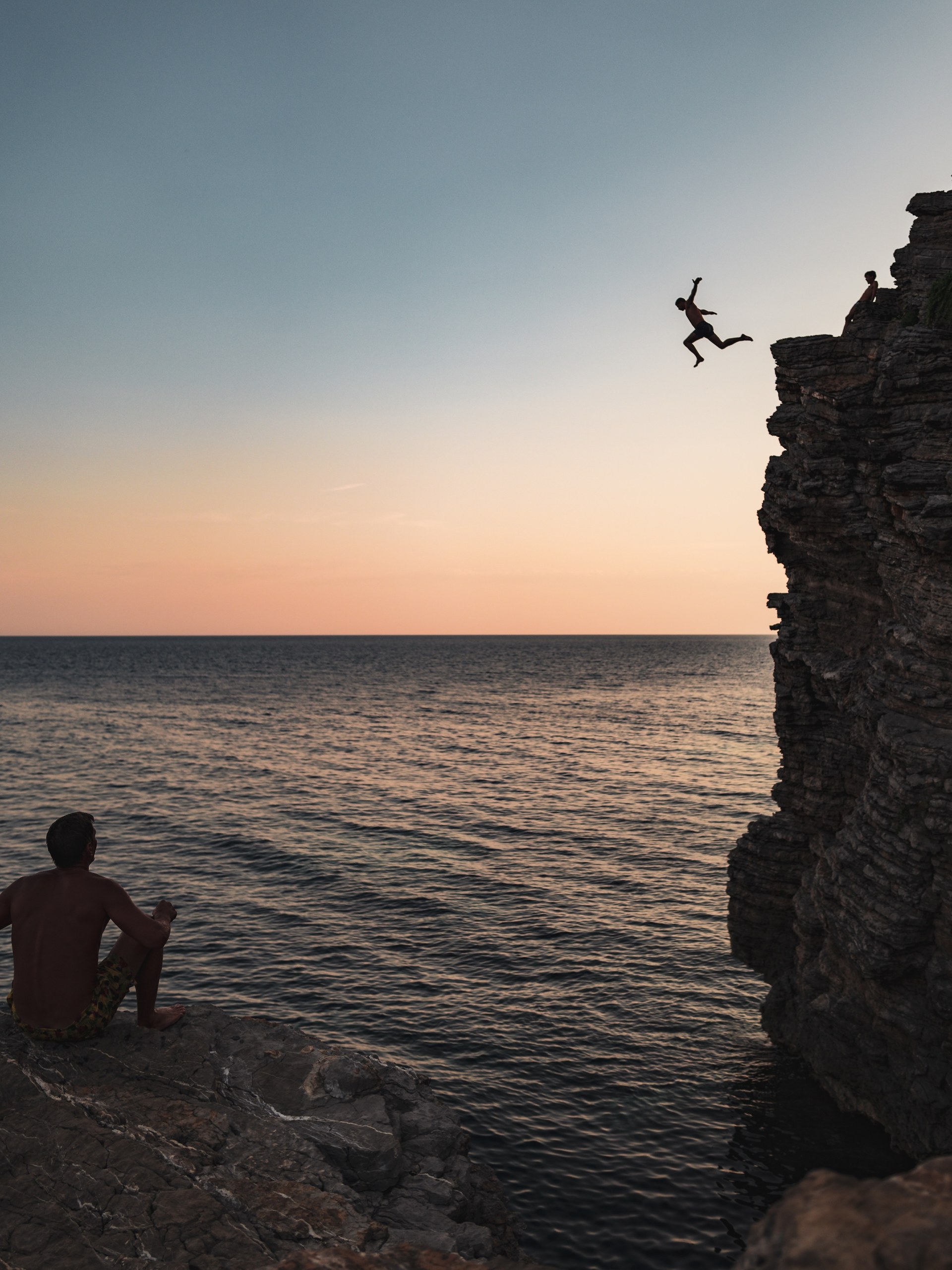 Person jumping off a cliff into the sea at sunset while another watches from the rocks, Budva, Montenegro