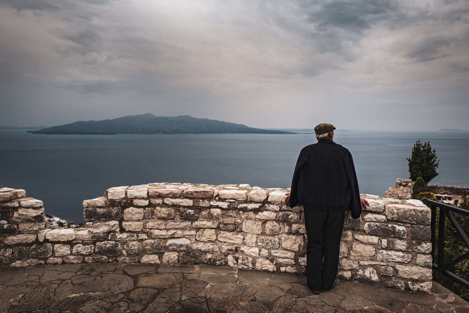 Elderly man standing at a stone wall looking out over the sea to an island, Sarandë, Albania