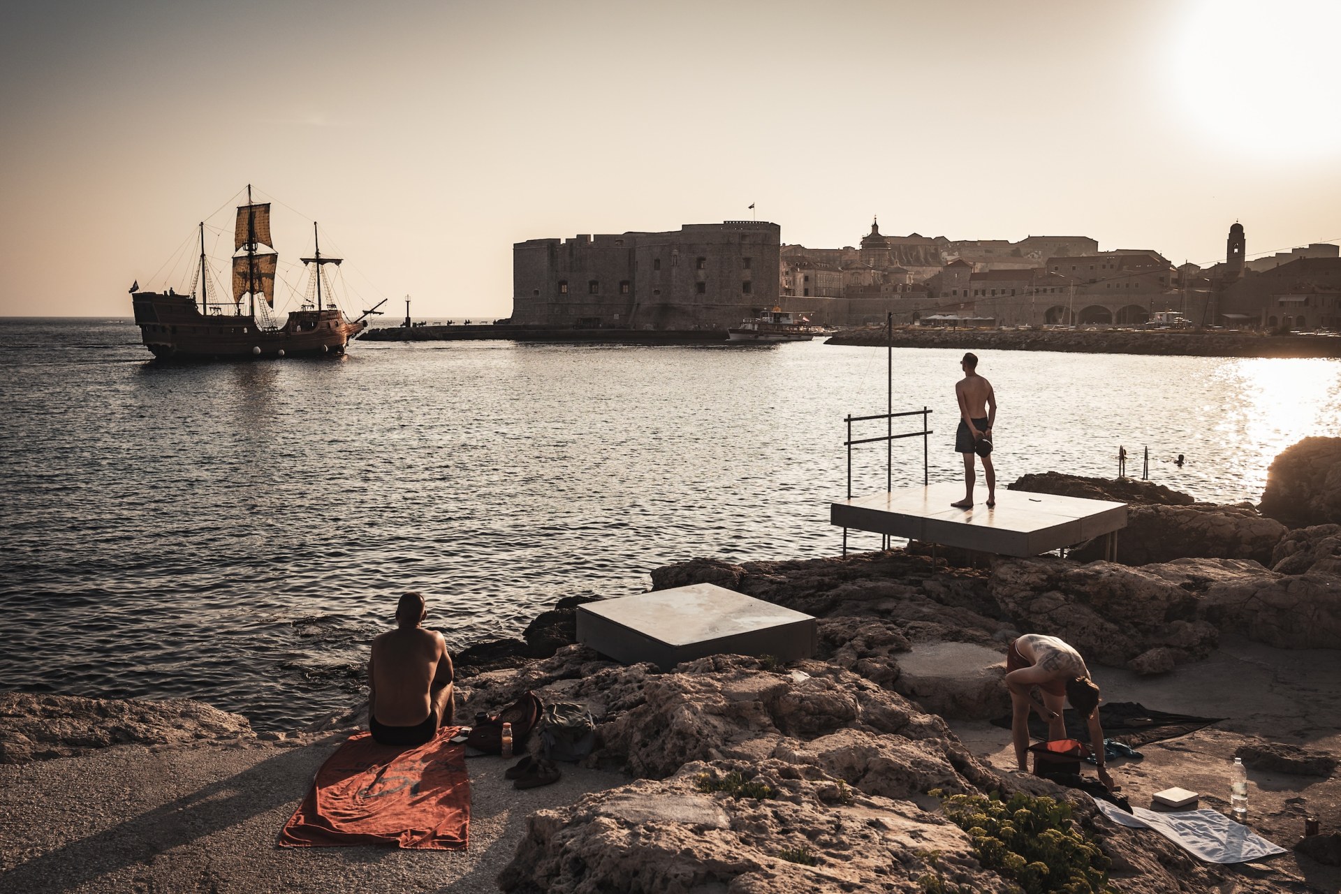 People on the rocks by the sea in front of an old fortress and a historic sailing ship, Dubrovnik, Croatia