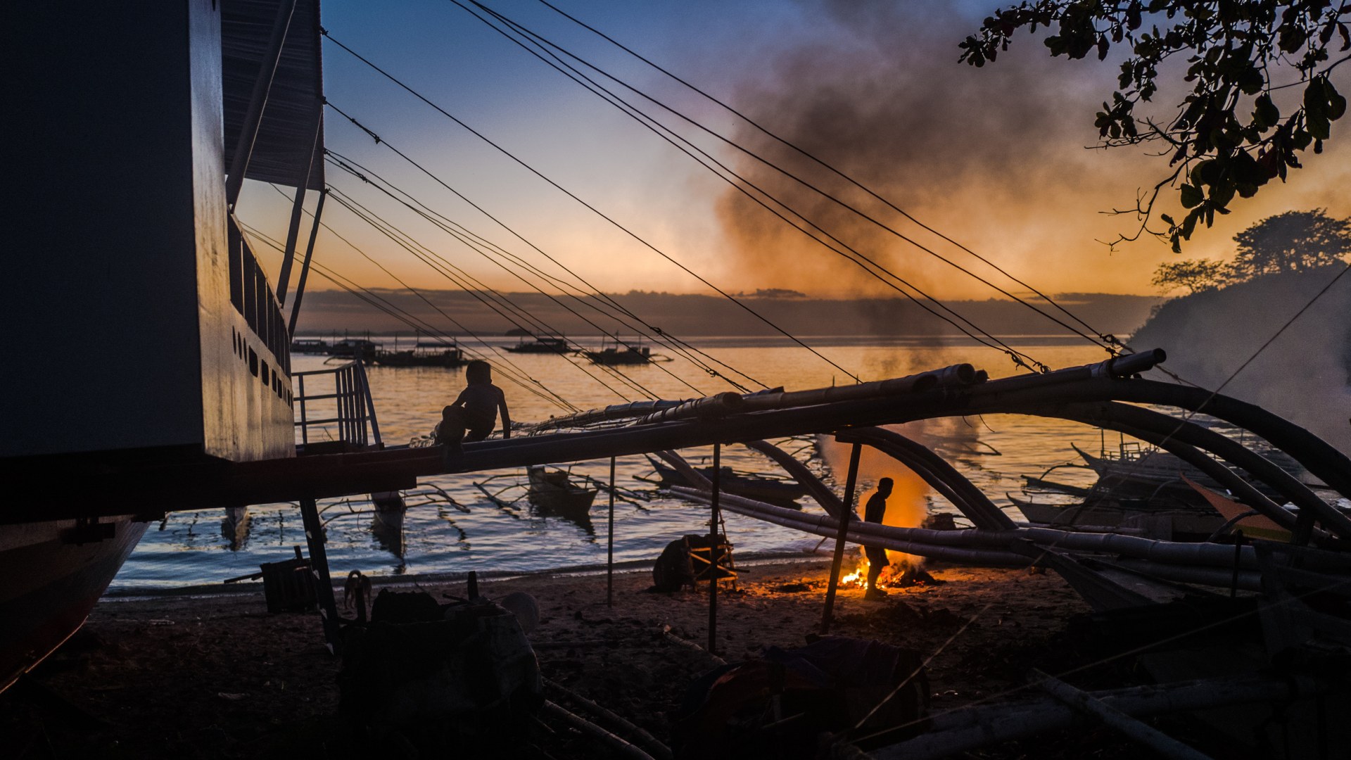 Child sitting on an outrigger boat at dusk while a man tends a fire on the beach, Malapascua, Philippines