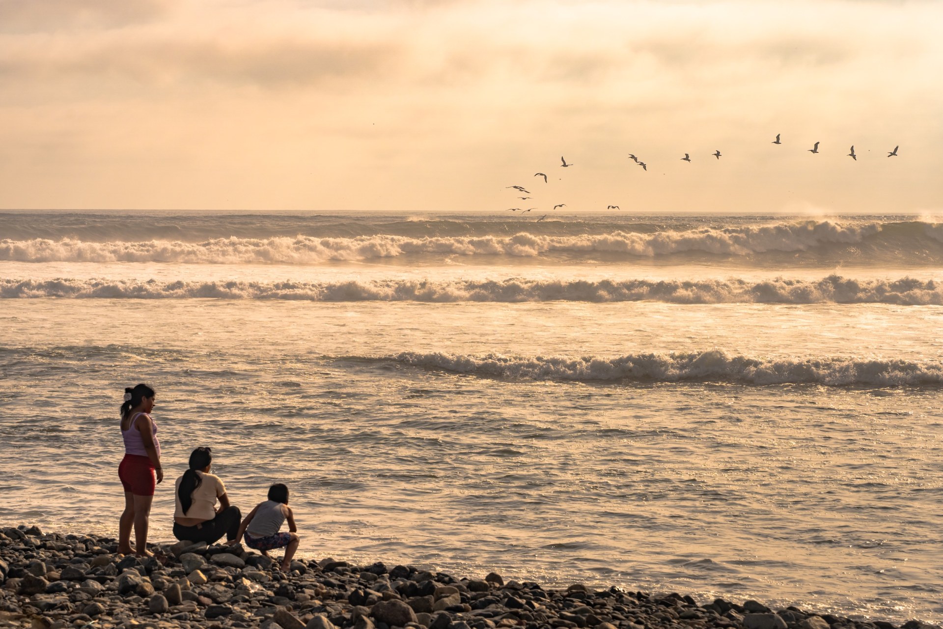 Three people on a pebble beach watching the surf as a flock of birds flies past at sunrise, Huanchaco, Peru