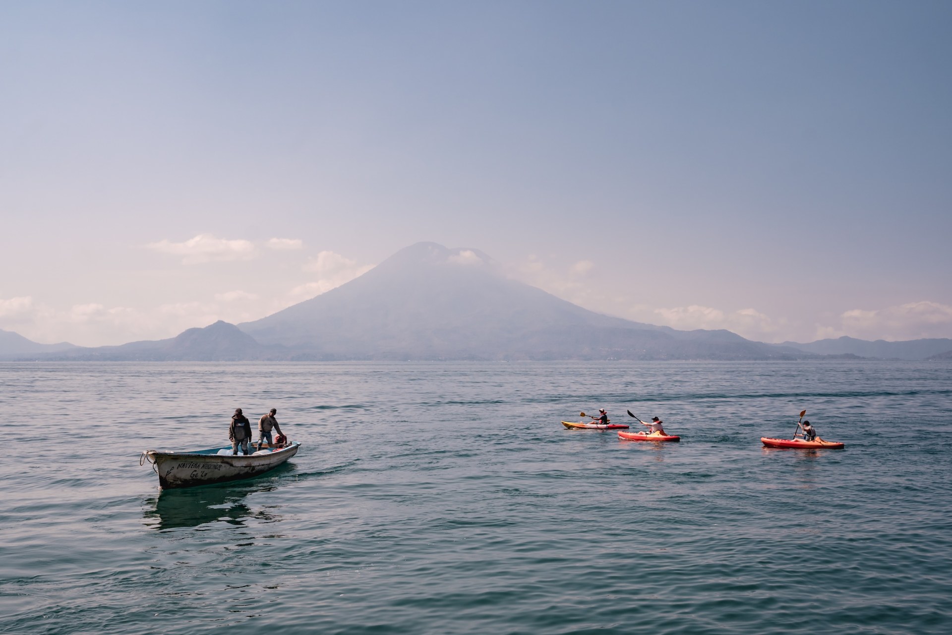 Two men in a wooden boat and three kayakers on Lake Atitlán beneath a volcano, Guatemala