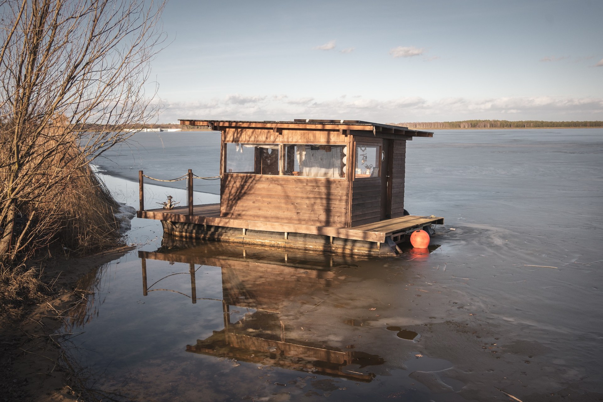 Floating wooden cabin on pontoons moored at the icy shore of a lake in Lusatia, Germany, reflected in the meltwater